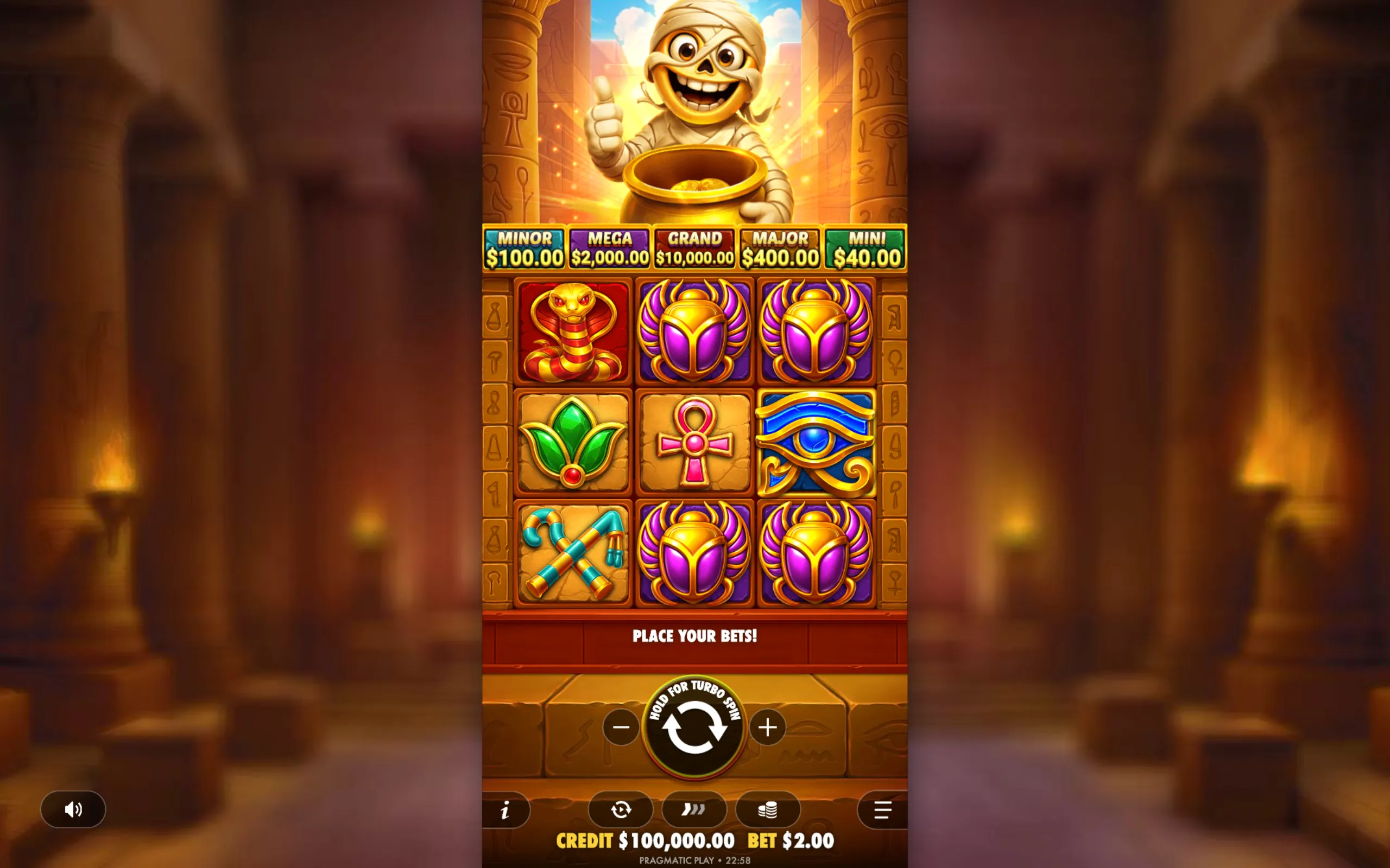 A colorful slot game interface featuring Egyptian-themed symbols like scarabs, ankh, eye of Horus, and a cheerful mummy holding a pot of gold coins, with jackpot tiers displayed above.